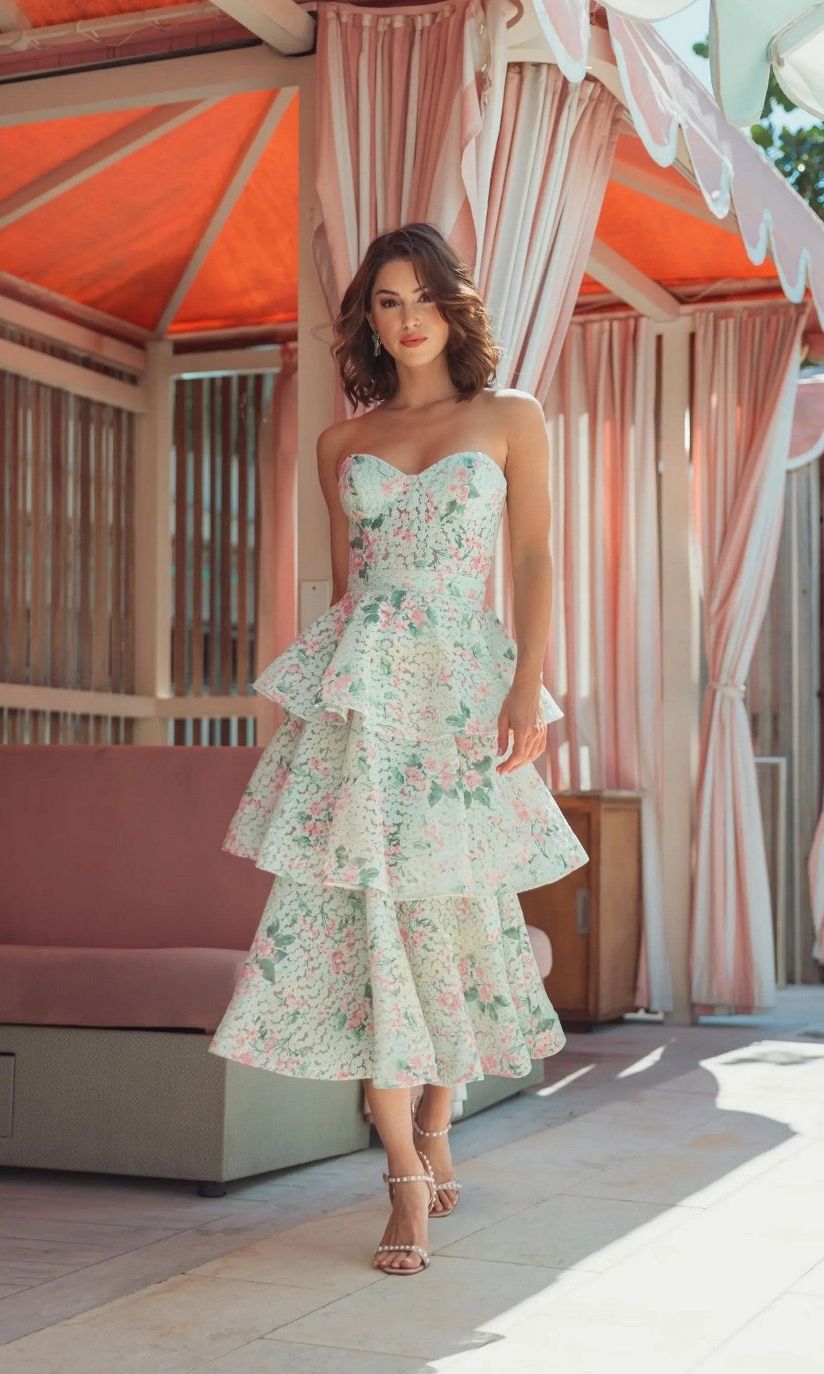 A woman stands outdoors beneath a canopy with pink curtains and wooden decor, wearing the Midi Wedding Guest Dress: Mac Duggal 56389 in Mint Multi—a strapless midi dress featuring tiered layers of floral-printed net—in a sunlit setting.